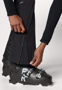 Cizmă de schi neagră cu accente metalice, catarame ajustabile și un logo alb. Mână ajustând manșeta unei perechi de pantaloni de zăpadă negri, cu material texturat.