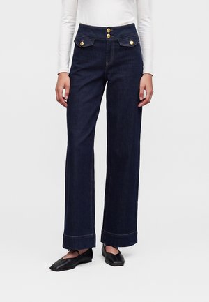 Personne portant un jean taille haute bleu foncé à jambes larges avec des détails de boutons dorés et des chaussures plates noires, debout devant un fond uni.