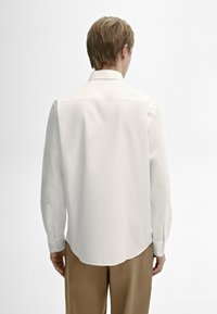 Chemise blanche à manches longues avec une texture douce, col pointu et poignets à un seul bouton, portée avec un pantalon marron clair. Design lisse et épuré.
