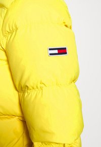 Žlutá péřová bunda s prošívanou texturou, s malou značkovou nášivkou v červené, bílé a modré barvě na levém rukávu.