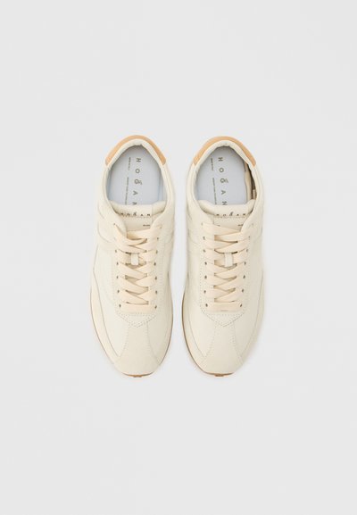Ζεύγος λευκών ανοιχτόχρωμων δερμάτινων αθλητικών παπουτσιών με κορδόνια, με μπεζ λεπτομέρειες στη φτέρνα, ραφές και επενδύσεις με λογότυπο σε απλό φόντο.