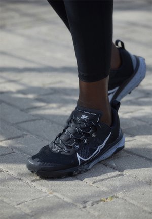 Gros plan d'une personne portant des chaussures de trail Nike Terra Kiger noires et des leggings noirs, debout sur une surface pavée.
