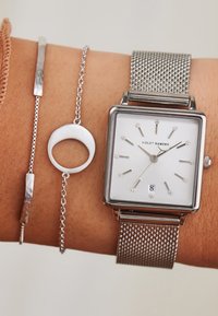 Vierkante zilveren horloge met mesh band, witte wijzerplaat, zilveren uurmarkeringen en datumweergave. Vergezeld van twee zilveren armbanden.