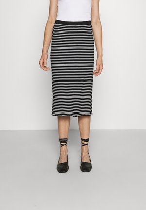 Femme portant une jupe noire et blanche rayée horizontalement jusqu'aux genoux, un haut blanc et des chaussures noires à lanières autour de la cheville, debout devant un fond uni.