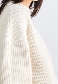 Pull tricoté couleur crème avec un motif texturé, des manches larges et un col montant, présentant une coupe décontractée et un matériau doux.