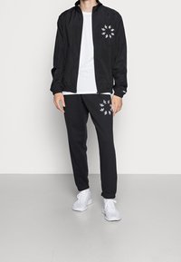 Svart lättviktsjacka med framfickor, vit t-shirt och svarta sweatpants med blommönster. Vita sportskor.