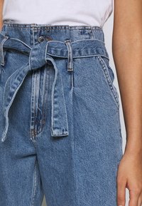 Vaaleansiniset farkushortsit, joissa on korkea vyötärö, edessä solmittava kangasvyö ja näkyvät ompelusaumat.