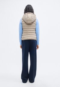 Μπεζ καπιτονέ γιλέκο με κουκούλα, με οριζόντιες ραφές. Φοριέται πάνω από ένα ανοιχτό μπλε πουλόβερ και συνδυάζεται με σκούρο παντελόνι φαρδύ στο κάτω μέρος.