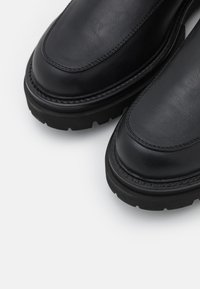Botas de tobillo de cuero negro con puntera redondeada, suela de goma resistente, acabado texturizado y costura visible a lo largo del borde.