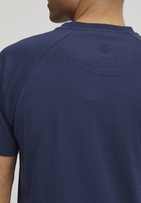 Temno modra majica iz mehkega bombaža, z raglan rokavi in tonirano embosirano logotipno podrobnostjo na hrbtni strani vratu.