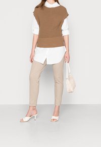 Pull sans manches marron tricoté sur une chemise blanche boutonnée, associé à un pantalon beige slim et des talons blancs ouverts, tenant un petit sac.