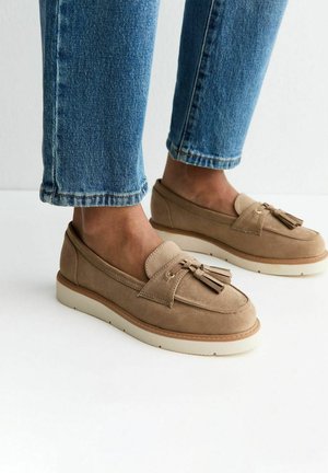 Pieds portant des mocassins en daim beige avec des pampilles, associés à un jean bleu raccourci, debout sur une surface blanche.