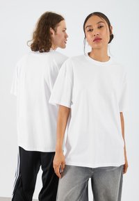 Valge unisex t-särk lühikeste varrukatega, ümmarguse kaeluse ja mugava lõikega. Ühel mudelil on sellega hallid püksid ja teisel mustad püksid.