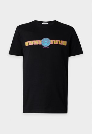 Man staat met handen in zakken, draagt een zwart T-shirt met goud- en blauw ontwerp, zwarte broek en zwarte schoenen met gouden neuzen.