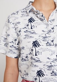 Košile s krátkým rukávem z bílé látky s tmavě modrým tropickým vzorem obsahujícím palmy a ostrovy. Klasický límec a zapínání na knoflíky.