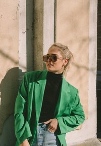 Blazer verde oversize su un dolcevita nero, abbinato a jeans a vita alta di un blu chiaro. Grande occhiali da sole marroni e una coda di cavallo bionda.