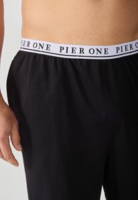 Svarte shorts laget av myk tøy med elastisk midjebånd som har hvit tekst "PIER ONE" og en lett glans.