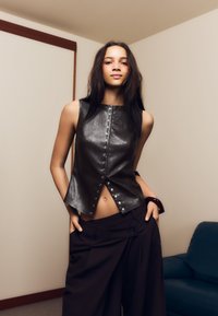 Crni kožni top bez rukava s kopčanjem na drukere sprijeda, u kombinaciji s oversized tamnocrnim hlačama. Glatka tekstura i usklađeni kroj.