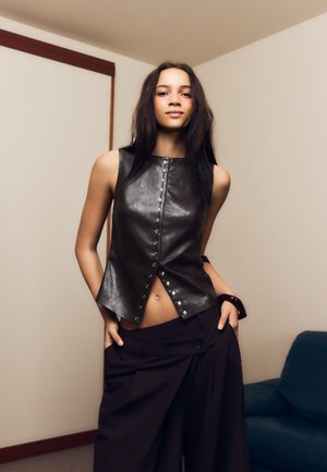 Top negru din piele fără mâneci, cu închidere frontală cu capse, asortat cu pantaloni largi de culoare maro închis. Textură netedă și croială cambrată.