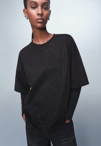 Black short-sleeve t-shirt featuring a subtle glitter pattern, crew neck design, relaxed fit, paired with a long-sleeve black top underneath.