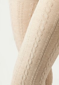 Knæhøje leggings i en lys beige nuance, med et struktureret kabelstrik mønster og blødt stof. Elastisk åbning for en tætsiddende pasform.