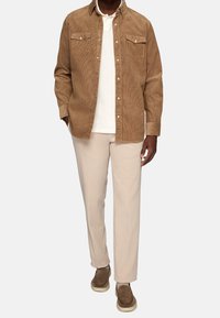 Bruine corduroy shirt met twee borstzakken, witte t-shirt eronder, lichtbeige broeken en bruine suède schoenen met rubberen zolen.
