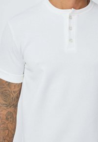Chemise henley à manches courtes blanche, dotée d'un tissu texturé, d'une patte de boutonnage à trois boutons et de poignets retroussés.