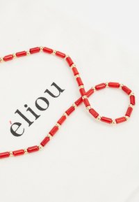 Piros gyöngy nyaklánc arany díszítéssel, henger alakú gyöngyökkel és sima felülettel, egy világos háttéren elhelyezve, rajta az "élilou" felirattal.