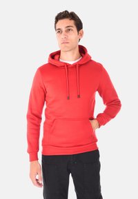 Uomo che indossa un hoodie rosso con tasca frontale e pantaloni neri, in piedi con una mano in tasca, che guarda di lato su uno sfondo bianco.
