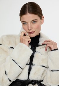 Kreativní kožešinová bunda v krémové barvě s černými akcenty, na zip, s širokými manžetami. Má páskový pas a načechrané prošívané části pro texturu.