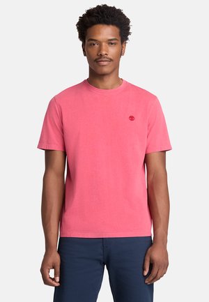 Mężczyzna stojący przodem, ubrany w gładki różowy t-shirt z krótkim rękawem i ciemnoniebieskie spodnie na białym tle.