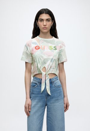 Kobieta z długimi ciemnymi włosami nosi kwiatowy krótki T-shirt z wiązaniem z przodu i logo "GUESS" oraz niebieskie wysokie jeansy, stojąc na białym tle.