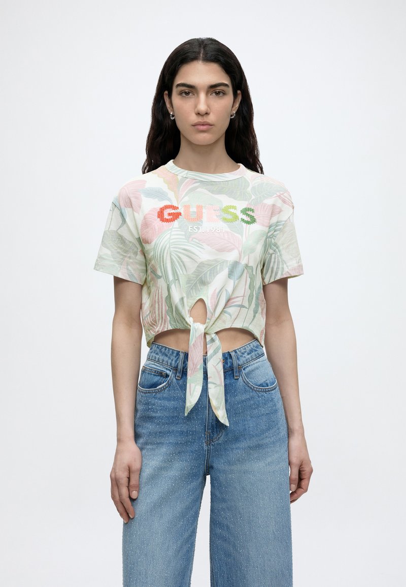 Γυναίκα με μακριά σκούρα μαλλιά φοράει κοντό φλοράλ μπλουζάκι με δέσιμο μπροστά και λογότυπο "GUESS" και ψηλόμεσο μπλε τζιν, στέκεται μπροστά σε λευκό φόντο.