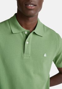 Zelené polo tričko s texturovanou tkaninou, s límečkem a dvoubarevným zapínáním, a malým vyšitým logem na prsou.