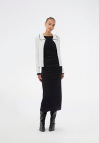 Μαύρο φόρεμα με ρυθμισμένο σχέδιο και λευκές βούλες, σε συνδυασμό με λευκή κάρντιγκ με μαύρη επένδυση. Το μοντέλο φοράει μαύρες μπότες μέχρι το γόνατο.