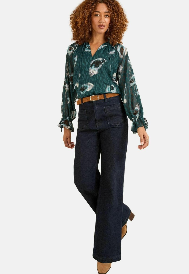 Blusa floreale verde con maniche a palloncino e scollatura a V profonda, abbinata a jeans a gamba larga blu scuro e una cintura marrone. Stivali con tacco marroni.