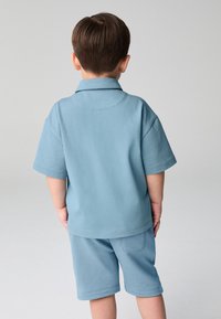 Ljusblå pikétröja med korta ärmar och krage, kombinerad med matchande shorts. Har en enfärgad design och mjukt, strukturerat tyg. Visar bakifrån.