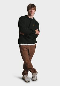 Sudadera negra con un pequeño logo de elefante amarillo, combinada con pantalones de pana marrones y zapatillas blancas. De pie con los brazos cruzados.
