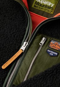 Zwarte fleecejas met groene voering, voorzien van zilveren ritsen en een oranje leren trekker, versierd met geborduurde patches op de borst.