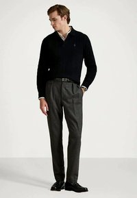 Man som bär svart långärmad tröja, vit skjorta, grå vikta byxor, svart bälte, klocka och svarta skor, står med en hand i fickan.