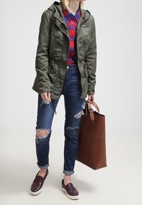 Olejově zelená bunda s kapucí, kostkovaná košile v červené a modré barvě, roztrhané tmavé džíny; hnědá taška a bordó slip-on boty. Hladké textury.