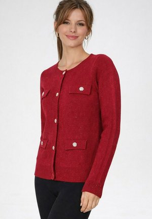 Femme portant un cardigan rouge ajusté avec des boutons argentés et quatre poches avant, associée à un pantalon noir, debout devant un fond blanc uni.