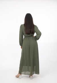 Vrouw met lang donker haar, gekleed in een olijfgroene maxi-jurk met lange mouwen en doorzichtige hoge hakken, staand met één hand op haar heup.