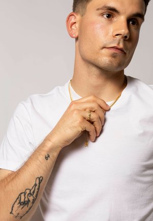 Guld smykker omfatter studs øreringe med en "A" accent, lagdelte halskæder og en tekstureret ring. Modellen bærer en enkel hvid t-shirt.