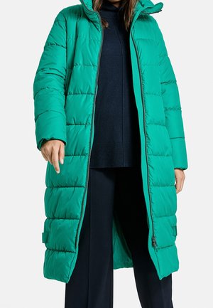 Long manteau matelassé bleu-vert avec capuche, entièrement dézippé, porté par-dessus un pull noir et un pantalon, visible des épaules aux genoux.