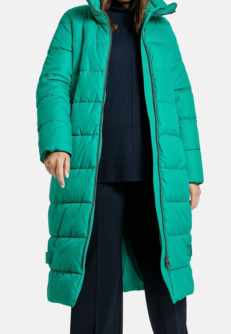 Long manteau matelassé bleu-vert avec capuche, entièrement dézippé, porté par-dessus un pull noir et un pantalon, visible des épaules aux genoux.