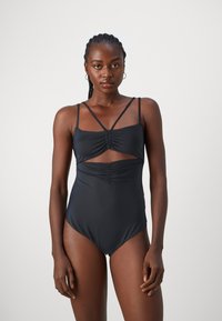 Svart én-delt badesuit med tynne, krossede stropper og samlingdetaljer ved brystet og midjeområdet. Glatt stoff og høyt utskårne benåpninger.