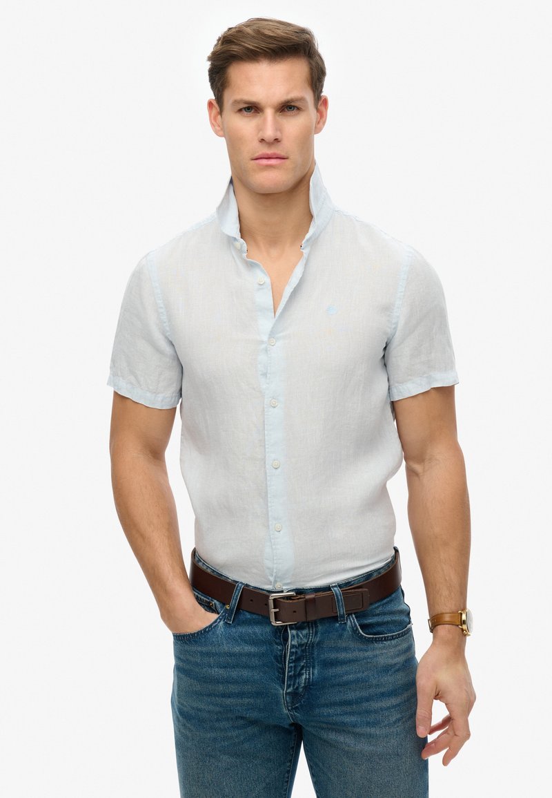 Jasnoniebieska lniana koszula z krótkim rękawem, zapinana na guziki, z kołnierzykiem, noszona z ciemnymi dżinsami i brązowym skórzanym paskiem.