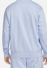 Osoba oblečená v světle modré mikině a odpovídajících kalhotách s vyšitým vzorem na pravé straně, zobrazená zezadu.