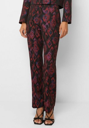 Pantalon à taille haute et jambe droite avec motif floral abstrait rouge, violet et bleu, porté avec des sandales à talons hauts noires à brides.
